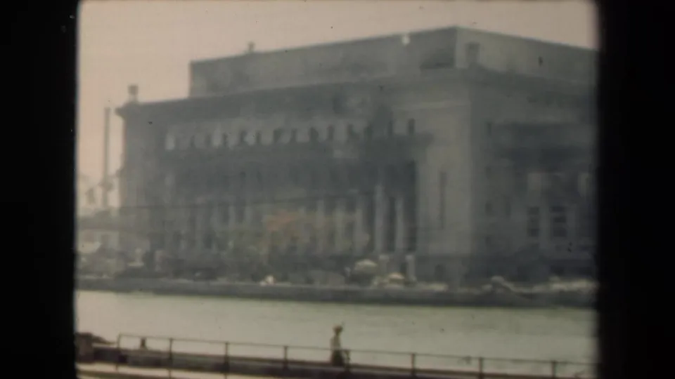 1944 Manila home-movie frame showing a large columned waterfront building across water, with a railing and distant pedestrian in front.
