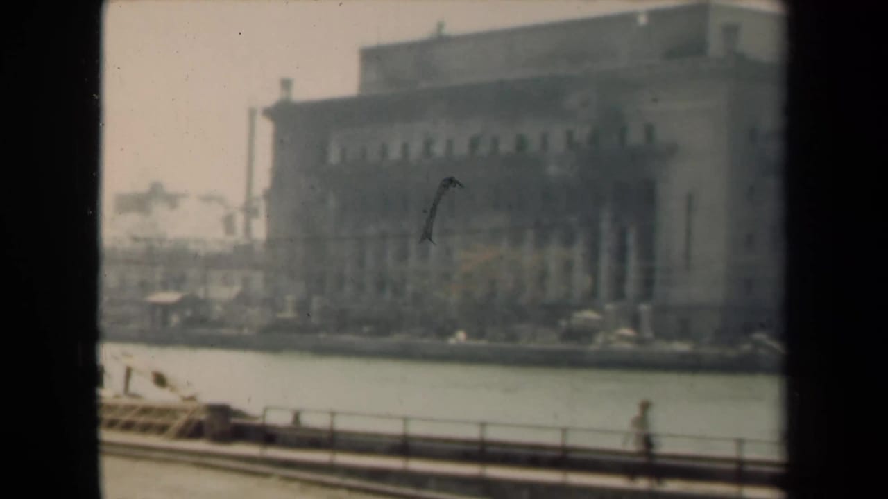 Similar waterfront view of the large building with water, railing, and a small walking figure.