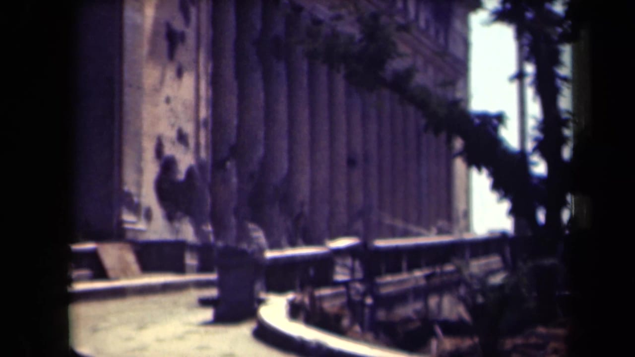 Side view of a damaged classical building beside a curved walkway and trees.