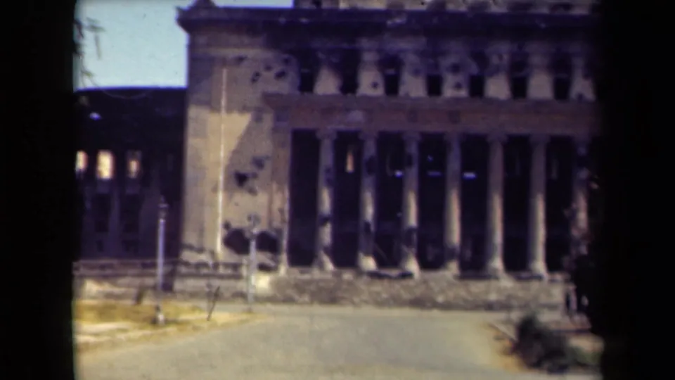 1944 Manila frame showing the front of a damaged columned building, with an empty road, dark interior openings, and trees at the edge.