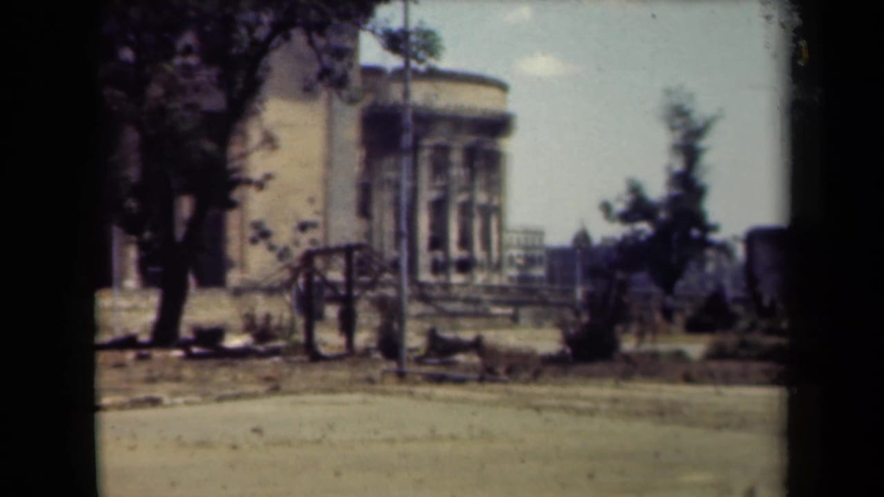 Distant damaged stone building beside a quiet street with trees and a lamppost.