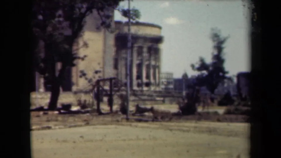 1944 Manila home-movie frame showing a damaged rounded stone building across a quiet street with trees, steps, and a lamppost.
