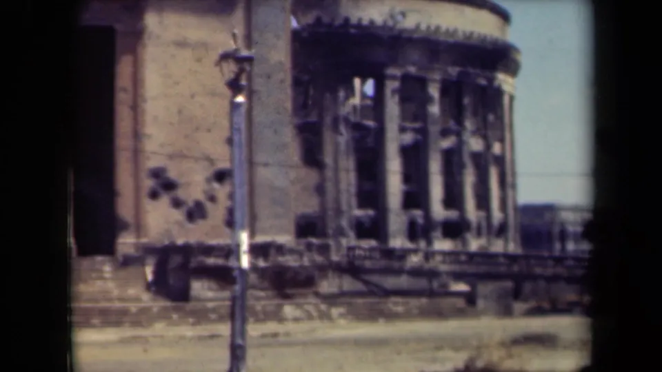 1944 Manila home-movie frame of a damaged stone building facade, with broken vertical window openings, a lamppost, steps, and an empty street.
