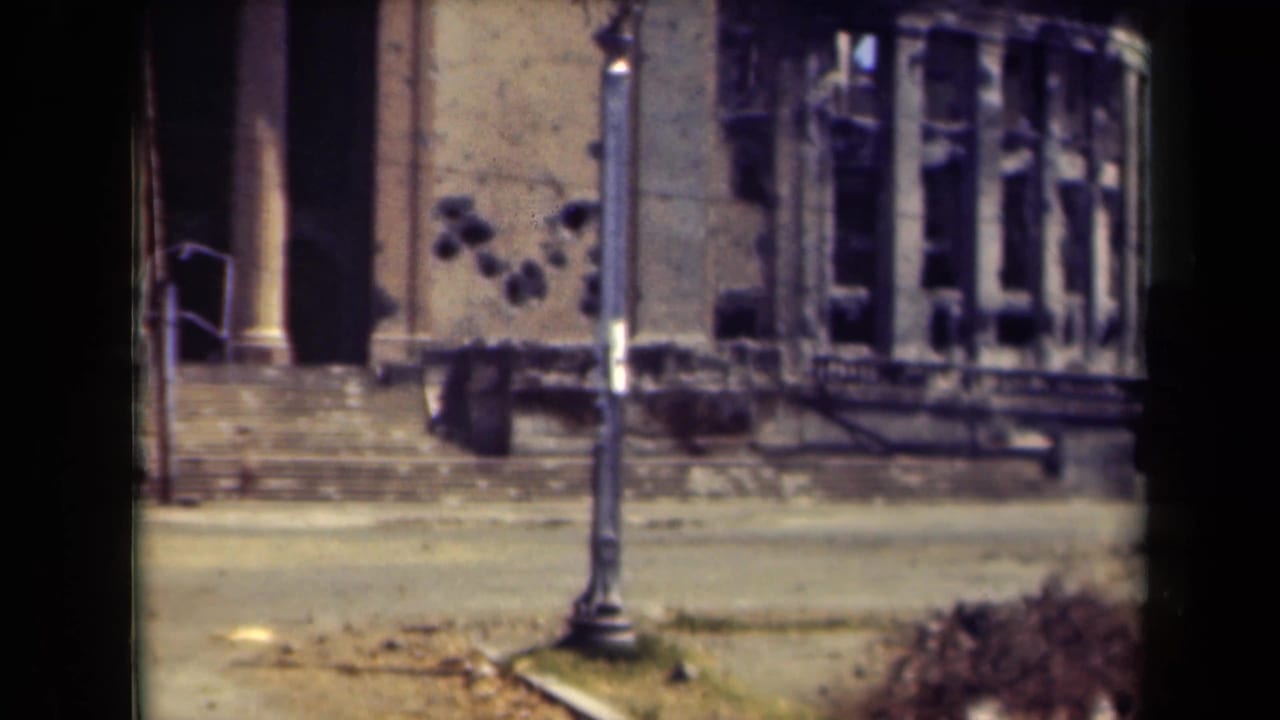 Damaged building facade with a lamppost, steps, roadway, shrubs, and dark window openings.