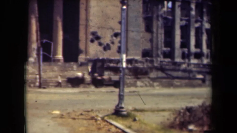 1944 Manila footage frames the damaged building closer, with shattered window openings, stone steps, roadway, and a lamppost before the facade.