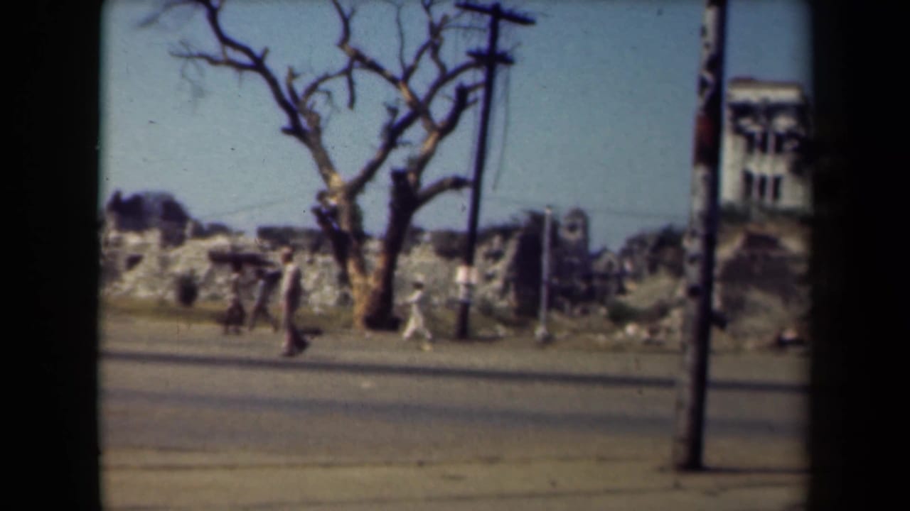 Pedestrians walk along a road beside rubble, a leafless tree and utility poles.