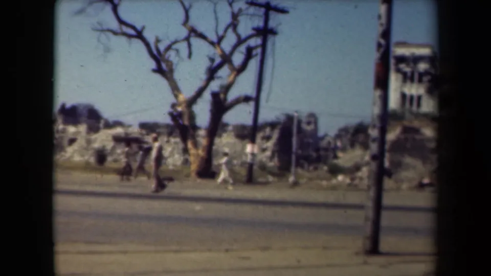 In Manila, Philippines, 1944, pedestrians move along a paved road beside rubble, damaged buildings, a bare tree and utility poles.