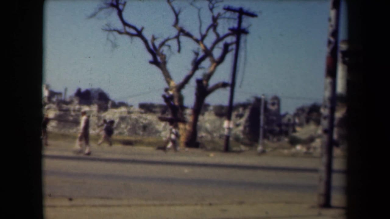 Several people walk past rubble and a bare tree along a broad road.