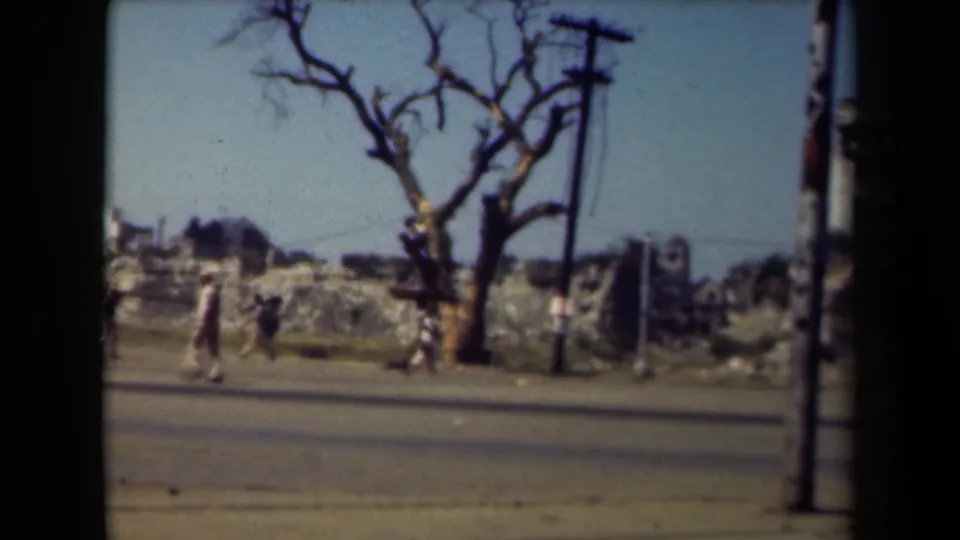 A 1944 Manila roadside view shows civilians walking past rubble, a leafless tree, utility poles and damaged structures.