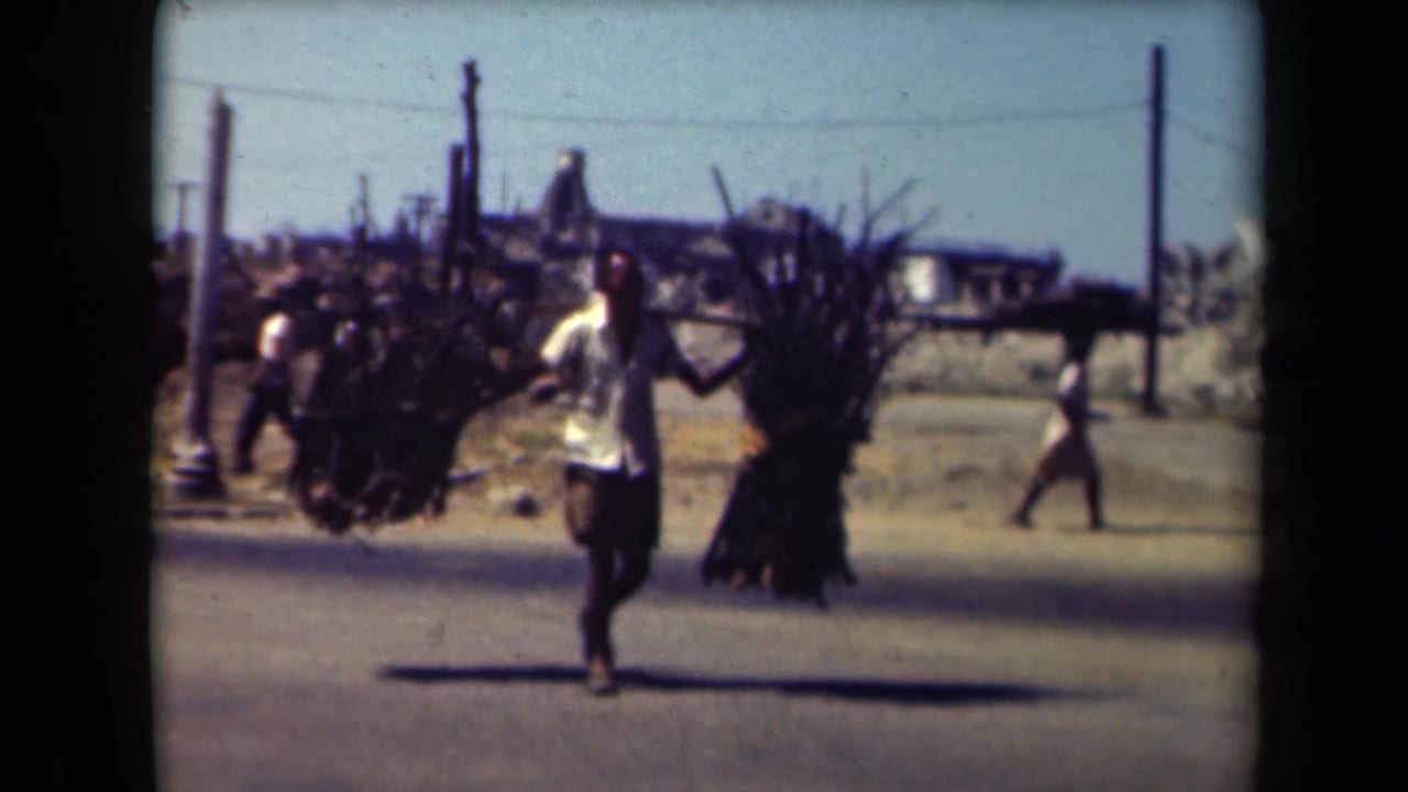 Man in a light shirt carries two large bundles along a sunny street as people pass behind him.