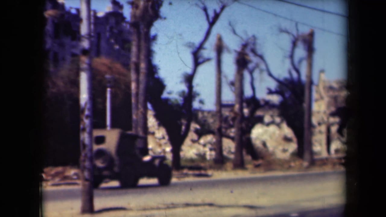 Boxy car driving past palms, utility poles, and damaged-looking roadside masonry.