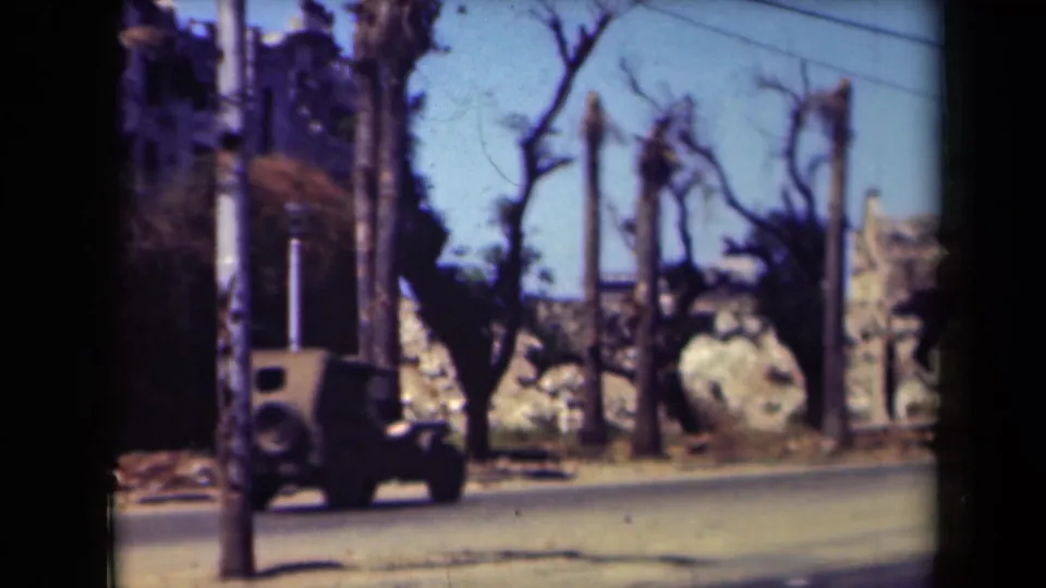 1944 Manila 8mm frame showing a boxy car on a sunlit street beside palms, bare trees, poles, and damaged-looking masonry.
