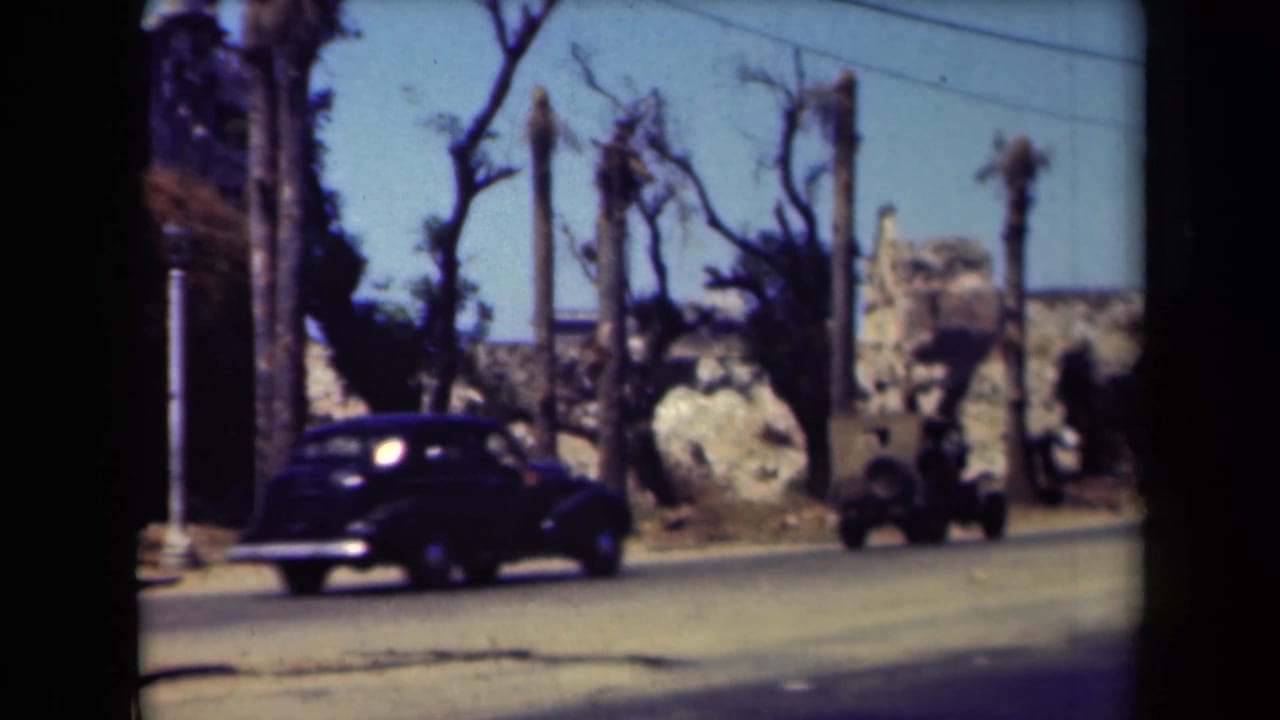 Dark classic car and jeep-like vehicle passing roadside palms and rough masonry.