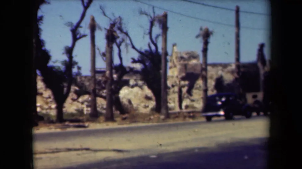 1944 Manila 8mm frame with a dark car at roadside, palm trunks, utility poles, overhead wires, and damaged-looking masonry in the background.