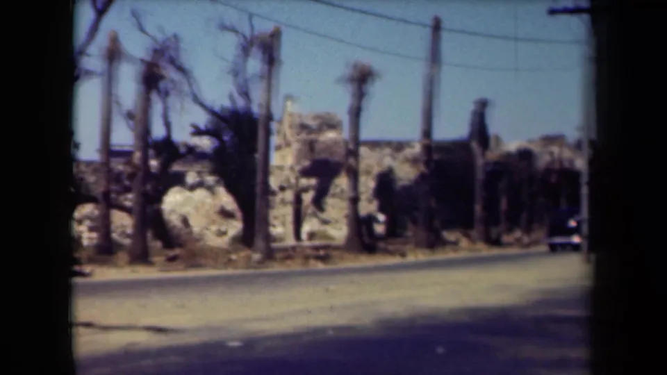 1944 Manila street frame showing a mostly clear road, palms, utility poles, wires, and damaged-looking roadside structures under bright sky.