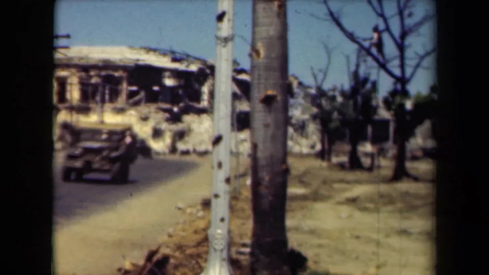 1944 Manila road scene with a military-style vehicle passing a heavily damaged building, bare trees, poles, and roadside debris.