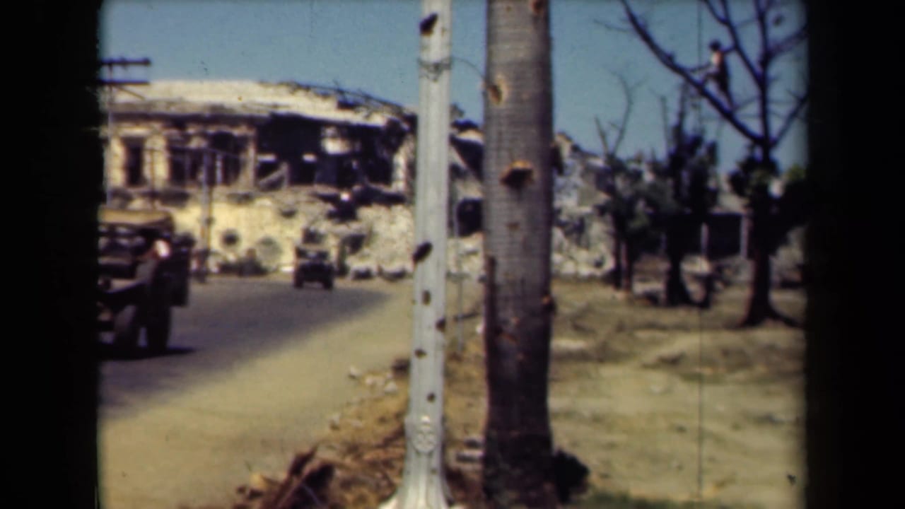 Damaged building beside a road with utility poles, trees, and a military vehicle at left.