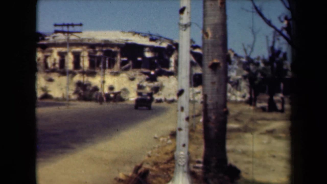 Ruined building and roadside poles seen across a street with a small vehicle in the distance.