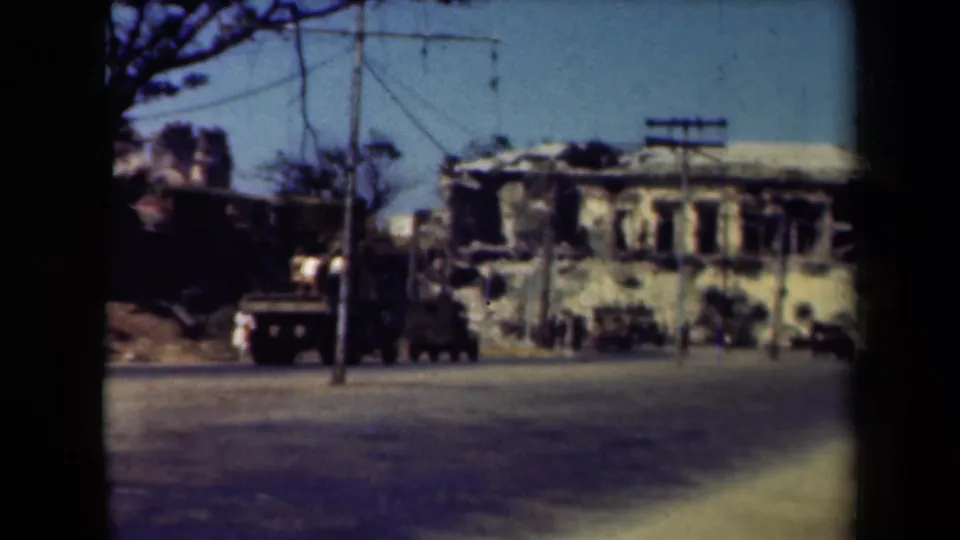 Wide 1940s Manila street scene showing damaged buildings, utility poles, roadside trees, and multiple military vehicles.