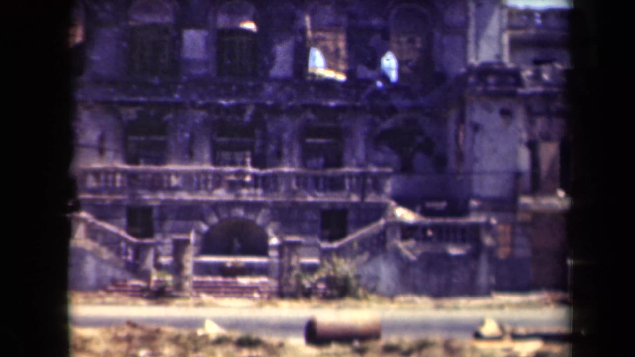 1944 Manila 8mm frame showing a war-damaged facade, broken windows, balcony railings, and debris along a sunlit road.