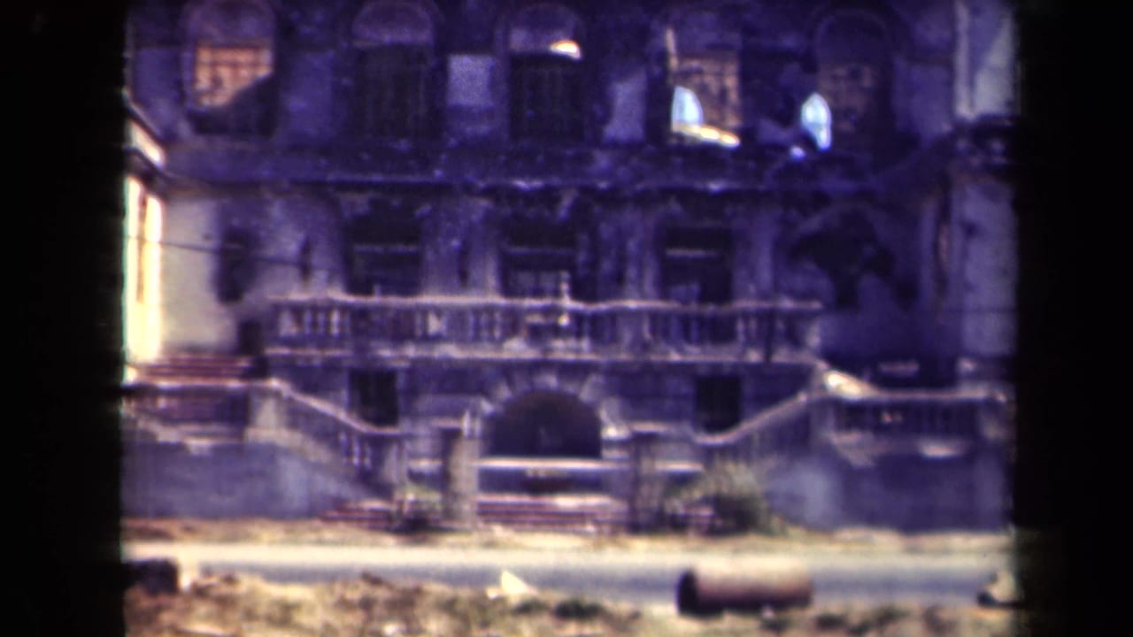 Ruined facade with balcony railings and a dark vehicle edge at right.