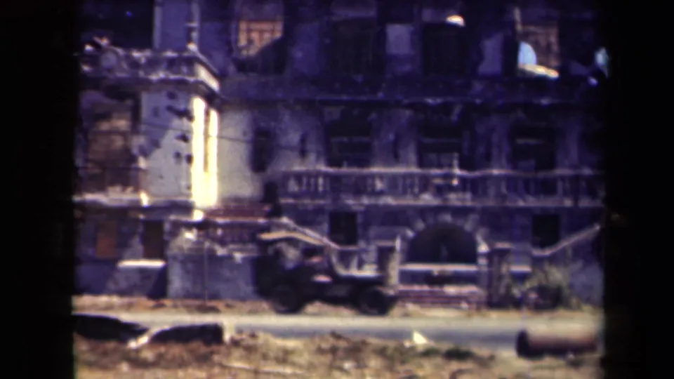 1944 Manila 8mm frame with a dark truck moving past a heavily damaged building facade and roadside rubble.