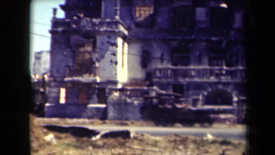 1944 Manila frame showing a dark truck passing a scarred building exterior with broken windows and debris near the road.