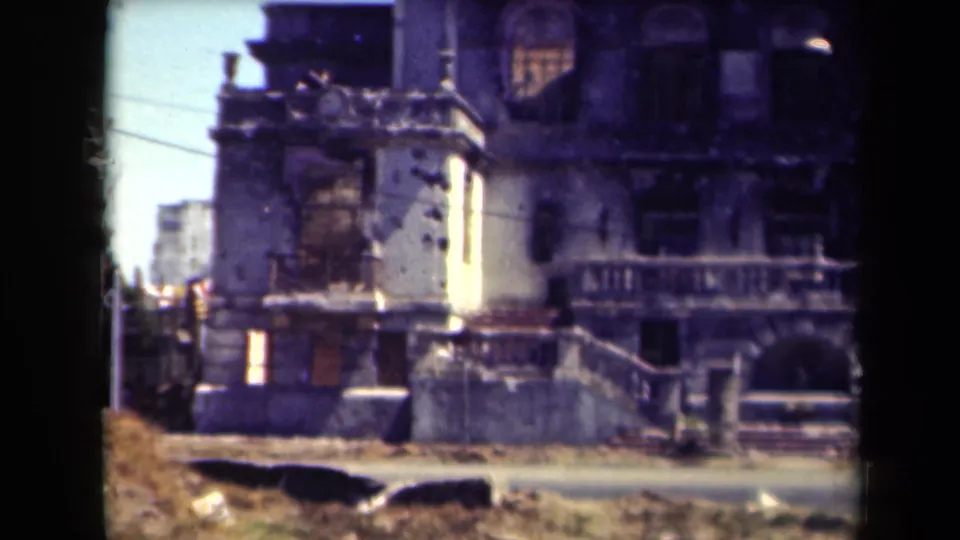 1944 Manila 8mm frame showing a war-damaged ornate building facade with shattered openings, stairwork, and street edge.