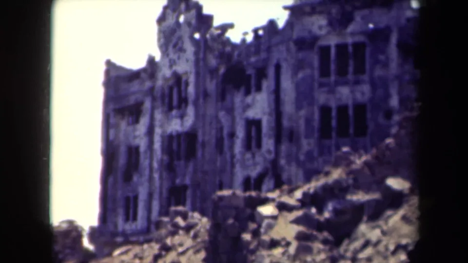 1944 Manila home-movie frame showing a damaged historic building facade with dark window openings and rubble in front.