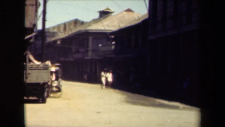 1940s Manila street view showing a parked truck, bicycle, pedestrians, utility poles, and older buildings under bright sunlight.