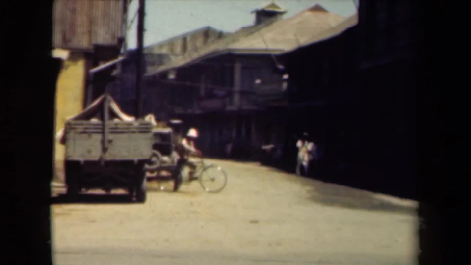 1940s Manila street frame with a truck, bicycle, pedestrians, and deep shade beneath older streetfront buildings.
