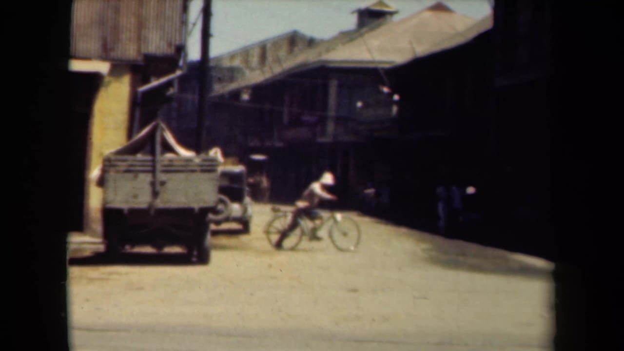 Bicyclist rides along a dusty street near a parked cart and low buildings.