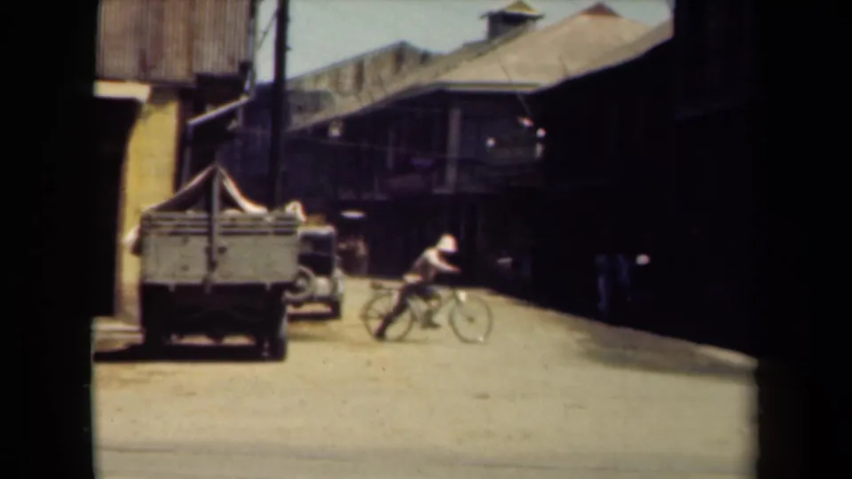 A bicyclist moves through a Manila street scene with a cart, shaded buildings, and a parked vehicle visible in faded 8mm footage.