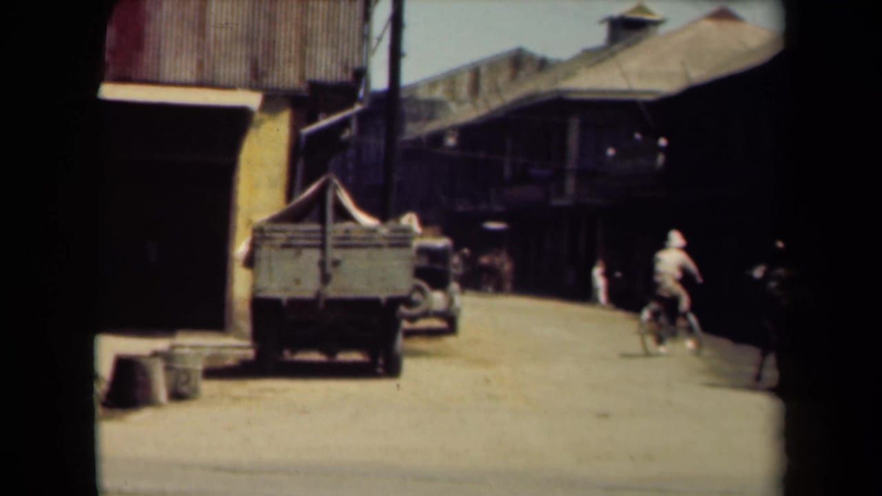 Bicyclist and pedestrians move past a cart, parked vehicle, and shaded streetfronts.