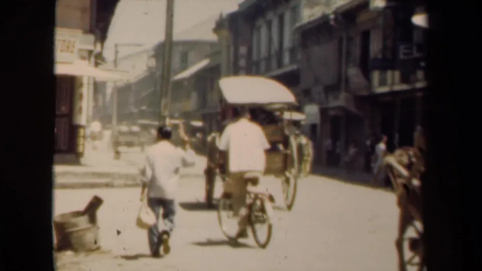 People walk and ride bicycles through a bright Manila street with storefronts, awnings, and weathered building facades.