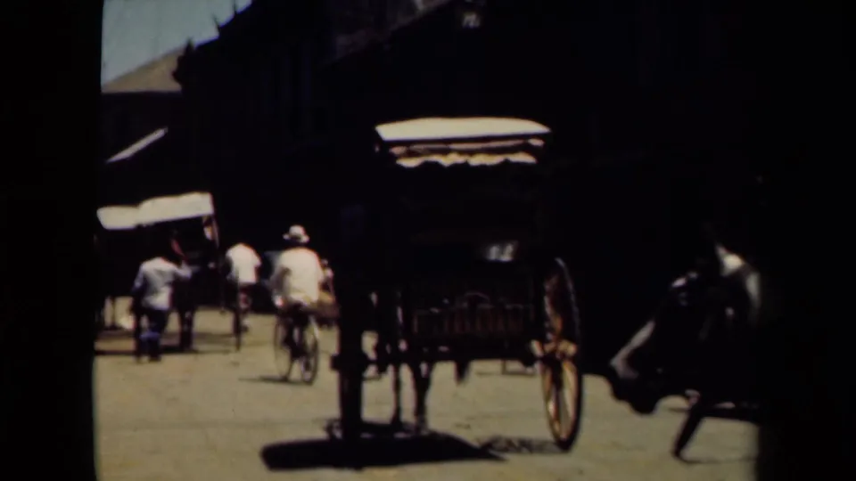 A horse-drawn carriage stands or moves near cyclists and pedestrians on a shaded Manila street in faded 8mm footage.