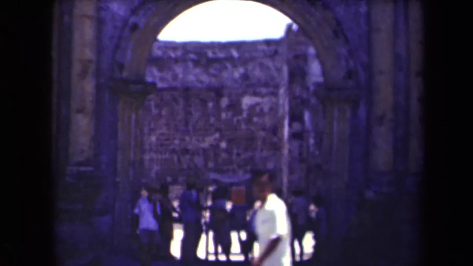 1940s 8mm frame of visitors near a weathered stone arch at a ruin site in Manila, with soft focus and heavy shadow.