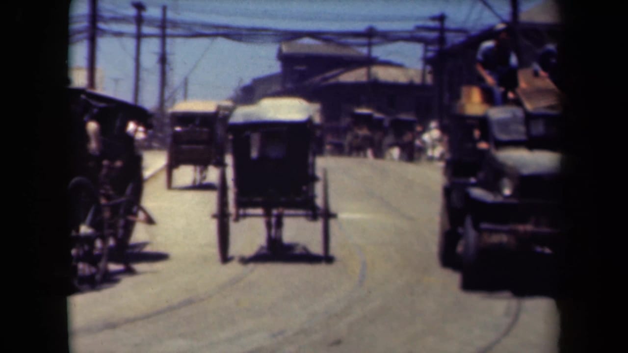 Carriage and motor vehicle pass along a road lined with pedestrians and utility wires.