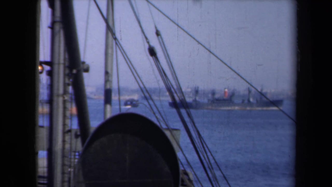 1946 New York Harbor view from a moving vessel, with rigging in the foreground and a distant cargo ship on hazy water.