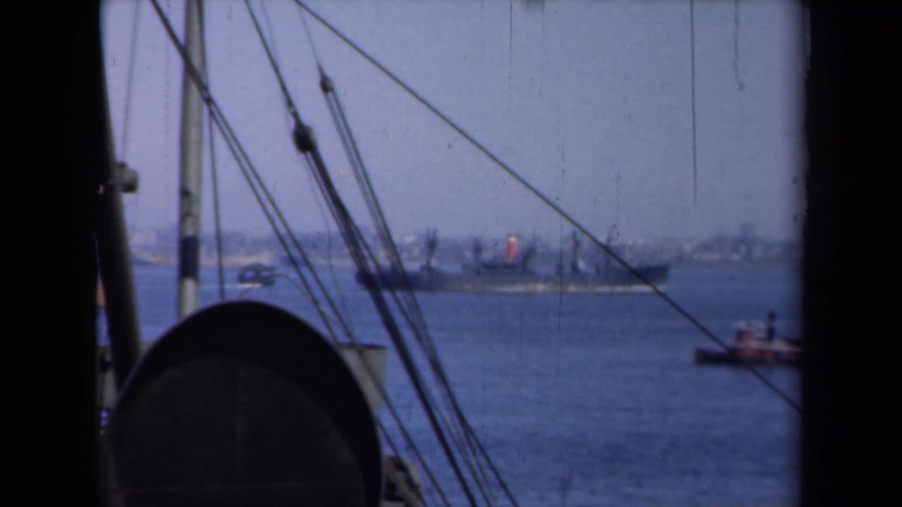 Cargo ship and small boat on harbor water, partly framed by rigging and dark vessel edges.