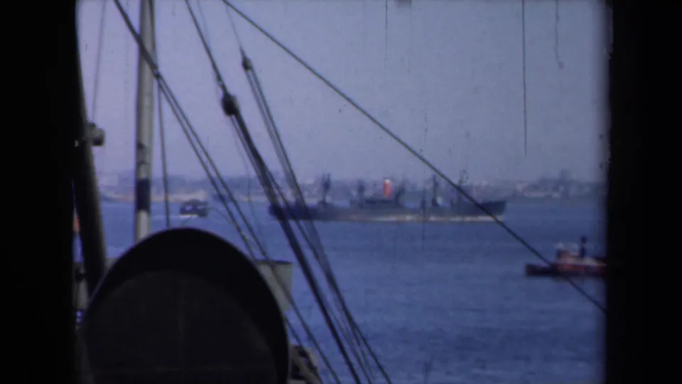 New York Harbor traffic appears beyond foreground rigging, with a cargo ship and small boat moving across the water.