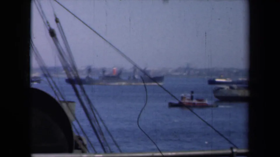1946 harbor view with a cargo ship, tugboat, and distant shoreline seen through foreground rigging from a moving vessel.