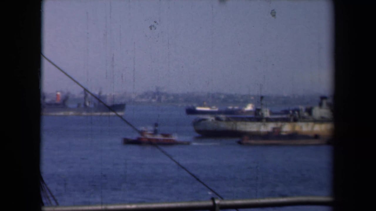 1946 New York harbor view with a large cargo ship, small tugboats, distant shoreline, and dark framing around the 8mm image.