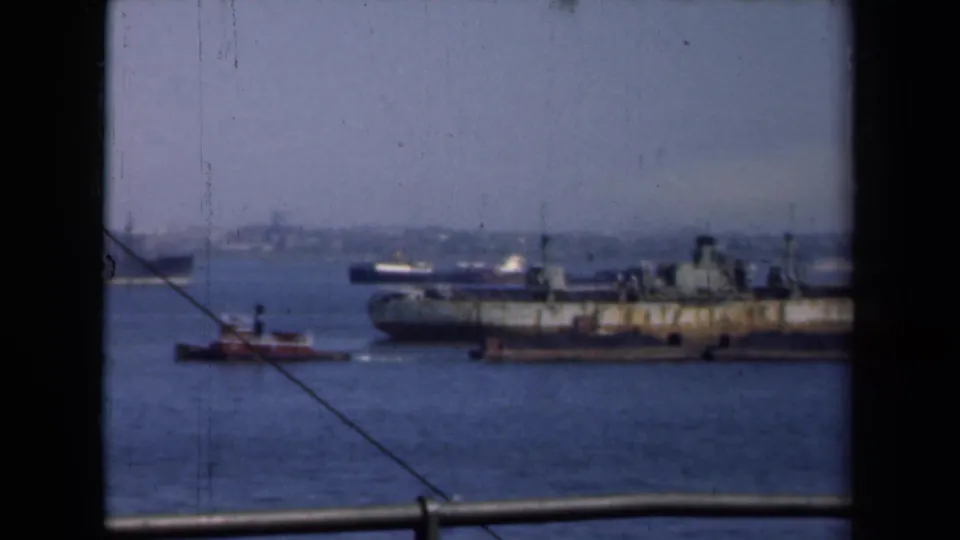 A large weathered cargo vessel passes through New York harbor in 1946, with small tugboats, distant shore, and window-like framing visible.