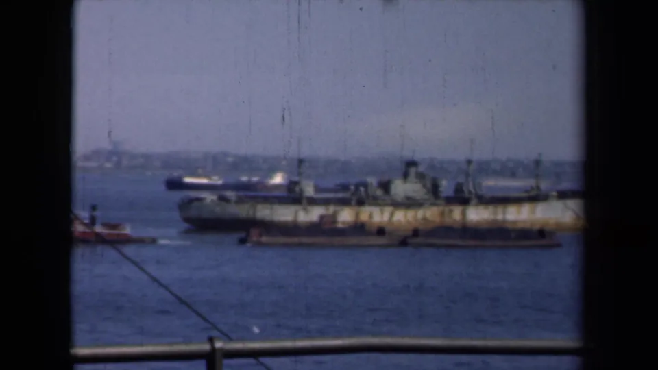 1946 New York harbor footage shows a weathered cargo ship moving past the camera, with tugboats, railing, and distant waterfront visible.