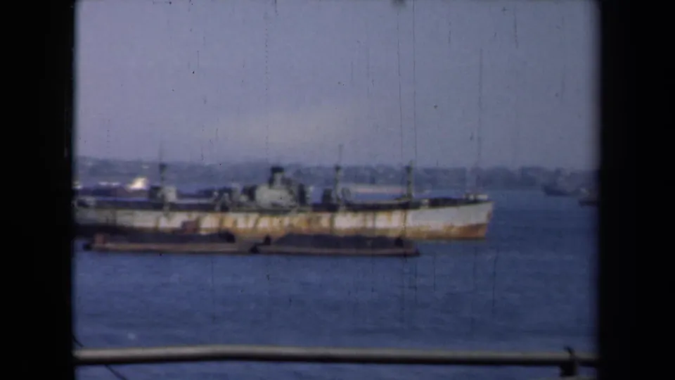 1946 New York harbor view of a rusty vessel on calm water, seen through dark vertical framing with a low shoreline beyond.