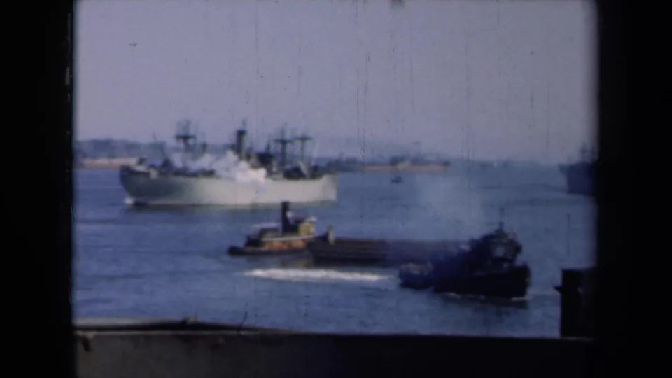 A tugboat moves across the foreground while a large ship sits or moves in the New York harbor background.