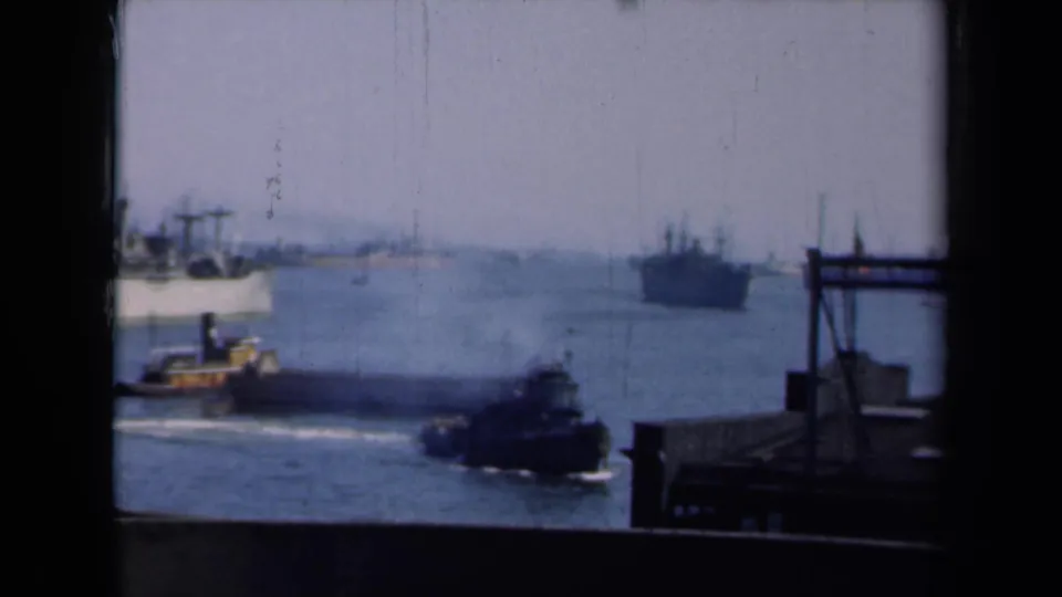 A New York harbor view shows a tugboat in the foreground, dock structure at right, and large vessels beyond.