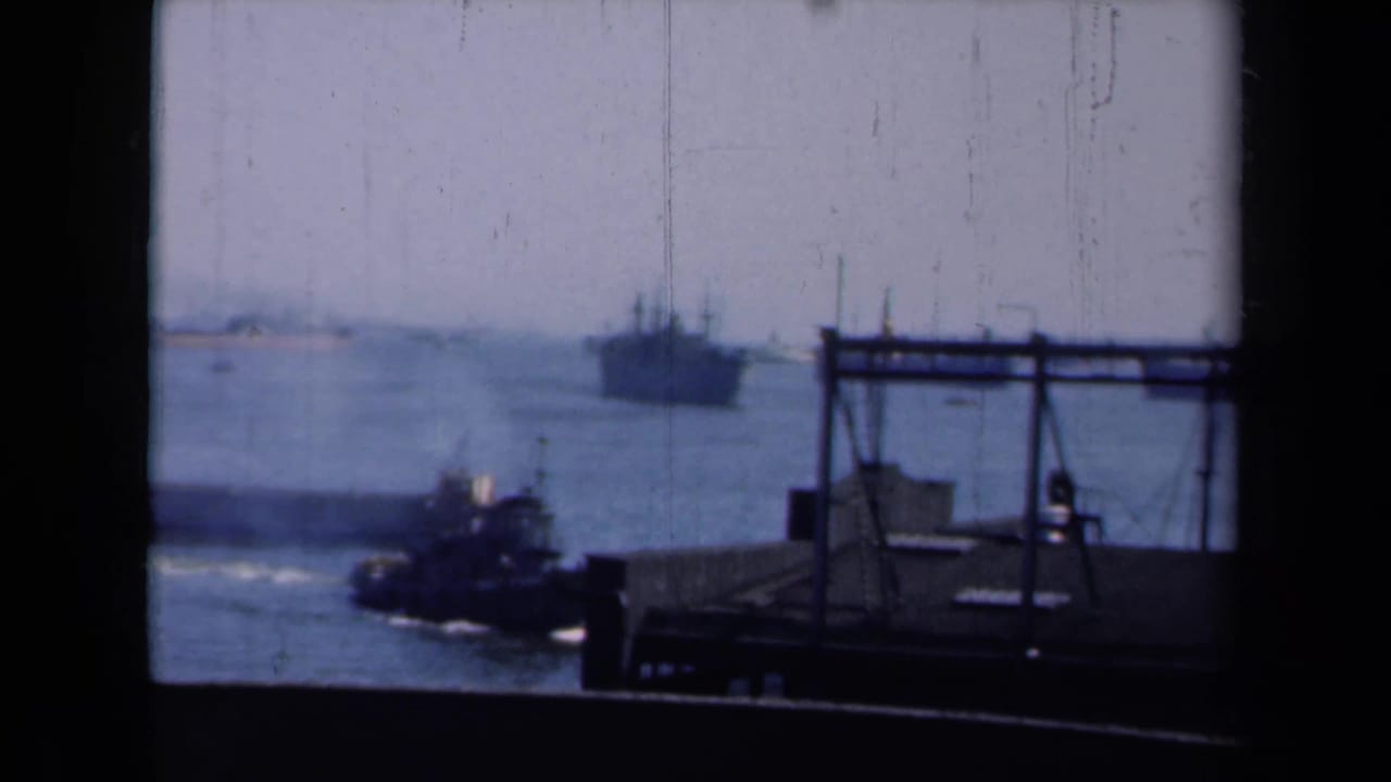 Tugboat passes near a pier while a large ship sits farther out in the harbor.