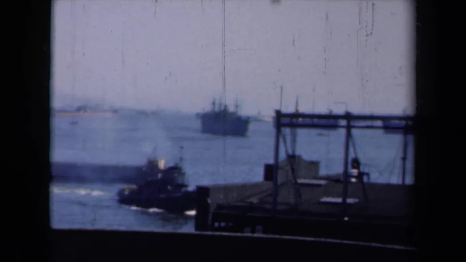 1946 New York harbor footage shows a tugboat near a dark pier structure, with a larger vessel in the distance across blue water.